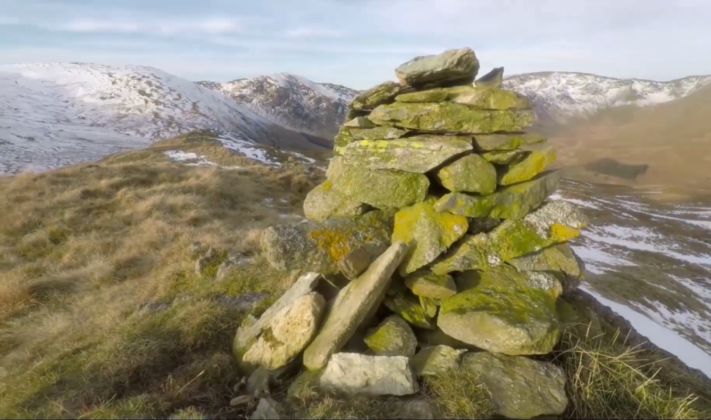 A moss-covered stone cairn stands on a hillside overlooking snowy mountains and a valley