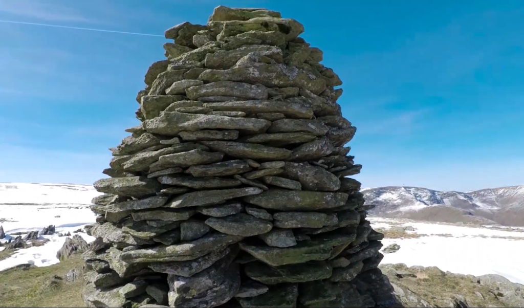 A tall stone cairn against a blue sky, with snowy hills in the distance